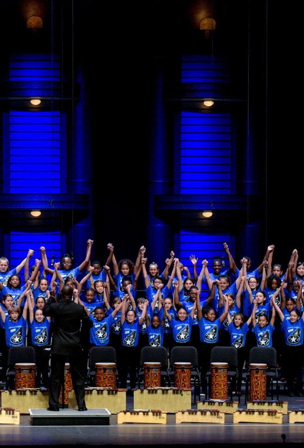 Kids performing on stage with a conductor. Their hands raised up.