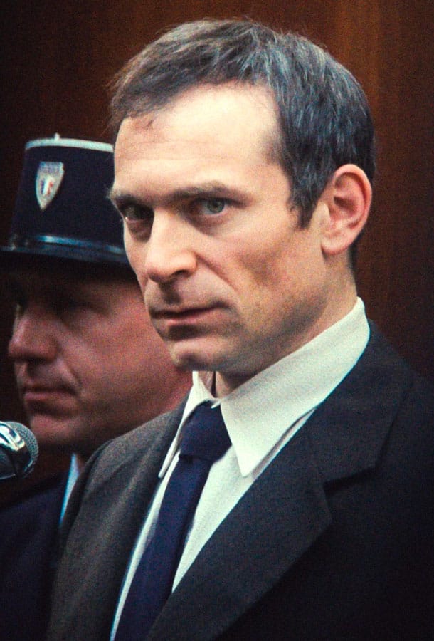 Mr. Goldman in a suit looking at the camera while being led by a police officer